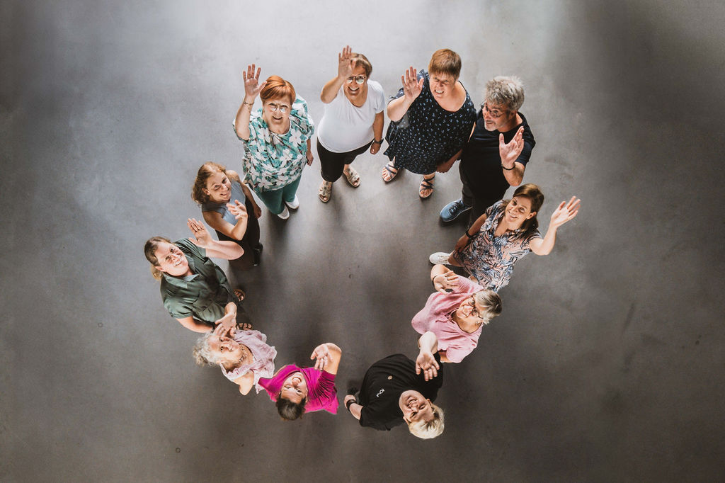 Groep deelnemers in een kring, van bovenaf gefotografeerd, zwaaiend naar de camera bij amba-amba in Desselgem