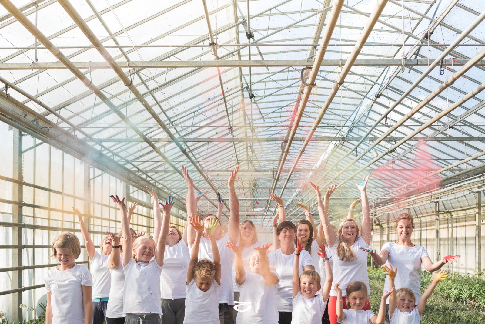 een groep mensen van kind tot volwassenen in een serre en ze gooien allemaal kleurpoeder in de lucht